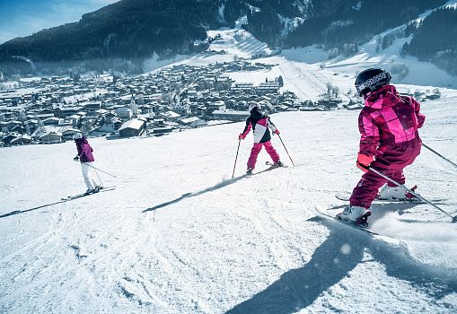 Drei Personen fahren gemeinsam auf einer verschneiten Piste mit Blick auf den Ort im Tal.