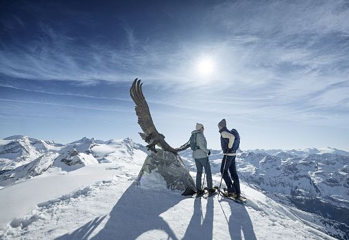 Zwei Personen stehen auf einem verschneiten Gipfel neben einer Adler-Skulptur mit beeindruckendem Bergpanorama.