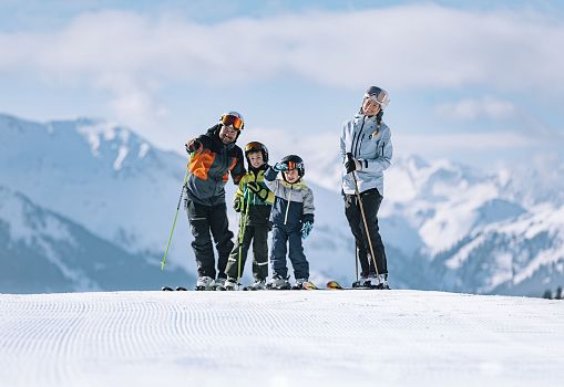 Family with two children smiling on a ski slope with snowy mountains in the background.