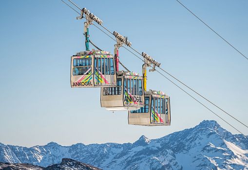 Drei bunte Gondeln einer Seilbahn vor verschneiten Berggipfeln.
