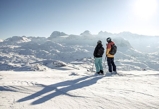 Zwei Skifahrer stehen auf einer sonnigen Piste vor beeindruckendem verschneitem Bergpanorama.