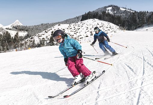 Zwei Skifahrer carven gemeinsam auf einer sonnigen Piste.