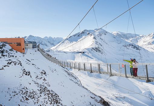 Zwei Personen stehen auf einer Hängebrücke in verschneiter Berglandschaft.