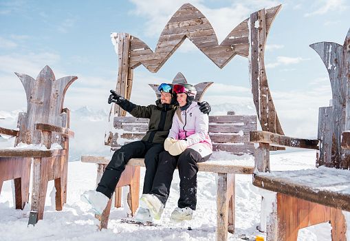 Zwei Personen sitzen auf einer großen Holzbank im Schnee vor winterlichem Bergpanorama.