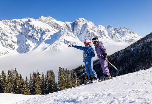 Zwei Skifahrer stehen auf einer sonnigen Piste und genießen das verschneite Bergpanorama.