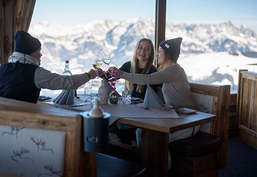 Drei Personen stoßen in einer Berghütte mit Blick auf verschneite Berge an.