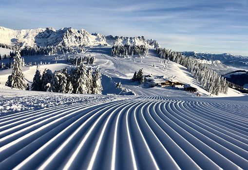View from a freshly groomed slope with a hut, snow-covered trees, additional slopes, and mountains in the background.