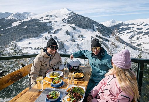 Three skiers enjoying traditional dishes on a sunny terrace with a snowy mountain view.