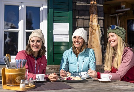 Three women happily enjoying coffee and cake on a sunny terrace in a ski resort.