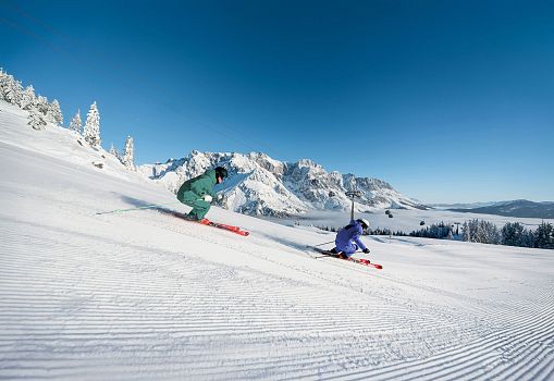 Two skiers carving on a freshly groomed slope with snowy mountain views.