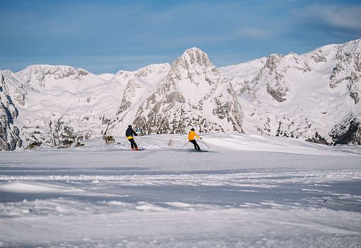 Two skiers on a wide snowy slope with impressive mountain scenery in the background.