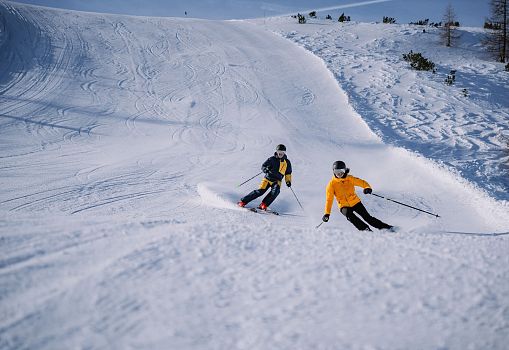 Two skiers carving together on a wide, snowy slope.