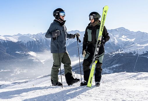 Zwei Skifahrer stehen auf einem verschneiten Gipfel mit Blick auf die Berge.