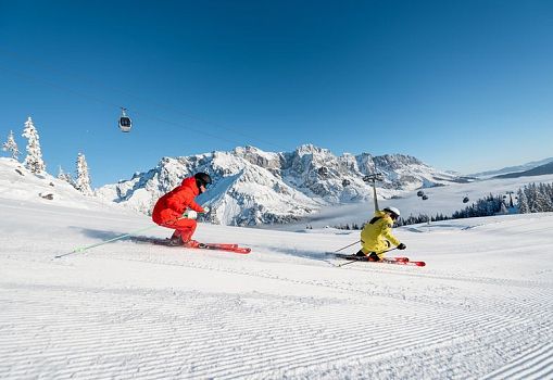 Two skiers carving side by side on a freshly groomed slope with mountain scenery and gondola lift.
