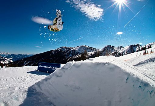 Snowboarder springt mit Backflip über einen Kicker im Snowpark bei Sonnenschein.