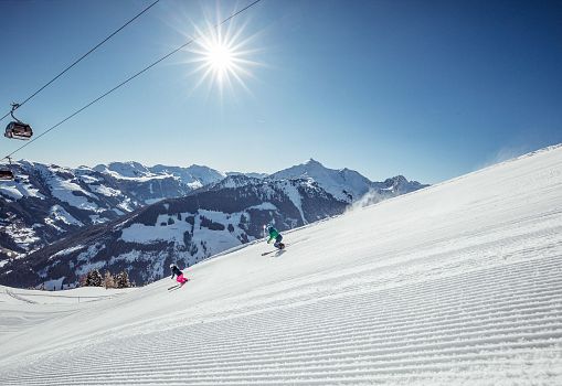 Two skiers on a freshly groomed slope with a mountain panorama in the background.