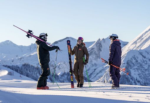 Drei Skifahrer stehen mit ihren Skiern auf einer sonnigen Piste vor verschneitem Bergpanorama.