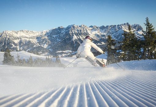 Female skier in white carving on a freshly groomed slope with a mountain backdrop.