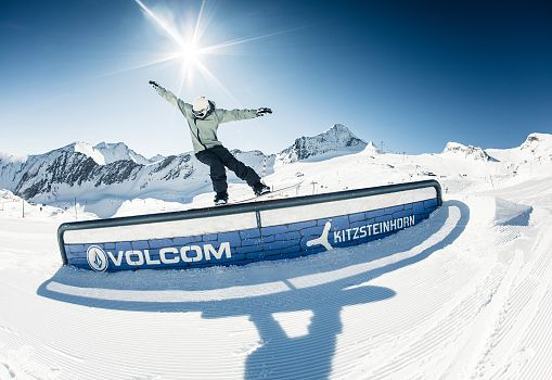 A snowboarder jumps over a rail in the Kitzsteinhorn Snowpark with snowy mountains in the background.