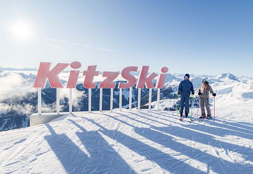 Two people standing in front of the KitzSki sign on a viewing platform with a snowy mountain panorama.