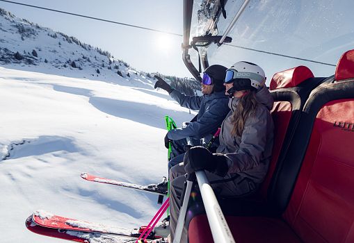 Zwei Skifahrer sitzen in einem Sessellift und genießen den Ausblick auf die verschneite Berglandschaft.