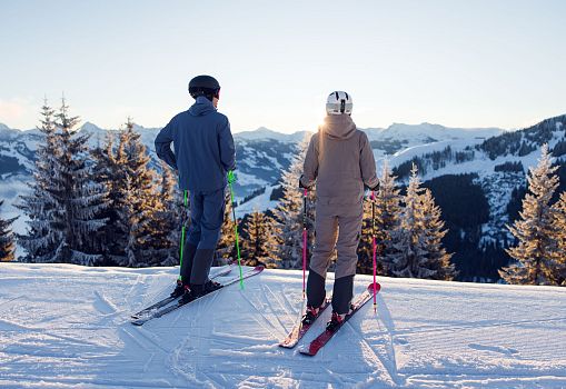 Zwei Skifahrer stehen auf einer verschneiten Piste und genießen den Ausblick auf die umliegenden Berge.