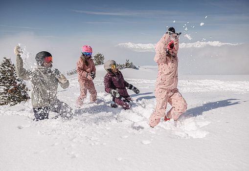 Eine Familie spielt gemeinsam im frischen Pulverschnee vor verschneiter Bergkulisse.