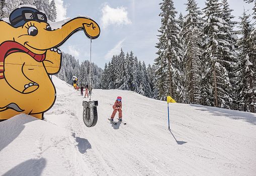 Ein Kind fährt auf einer verschneiten Funslope an einem Elefanten-Maskottchen vorbei.