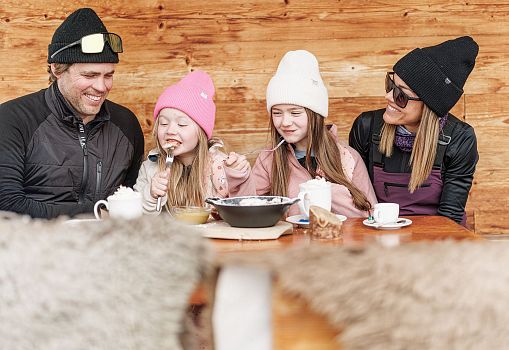 Eine Familie genießt gemeinsam Kaiserschmarrn und warme Getränke auf einer Berghütte.