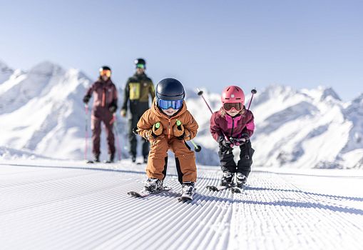 Two children ski down the slope while their parents watch in the background.