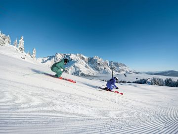 Two skiers carving on a freshly groomed slope with snowy mountain views.