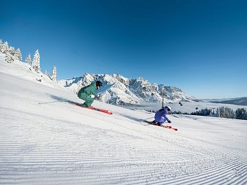 Zwei Skifahrer carven auf einer frisch präparierten Piste vor verschneitem Bergpanorama.